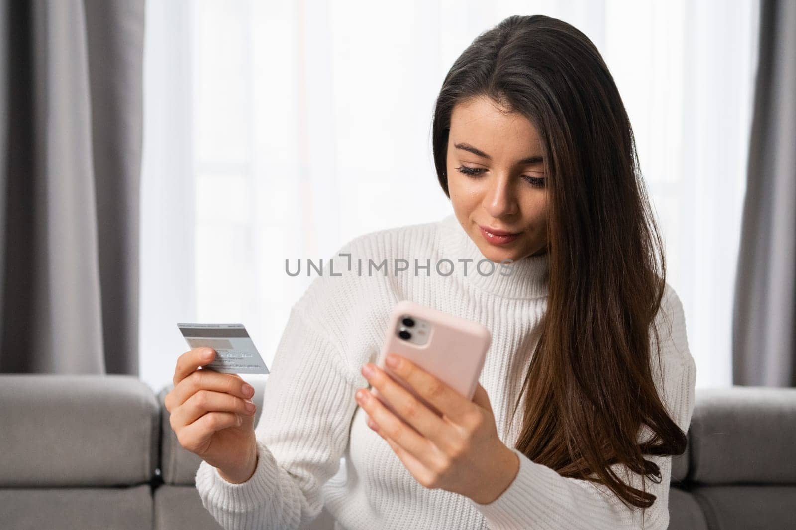 A caucasian woman with long dark hair pays by card using her mobile phone at home with a modern interior