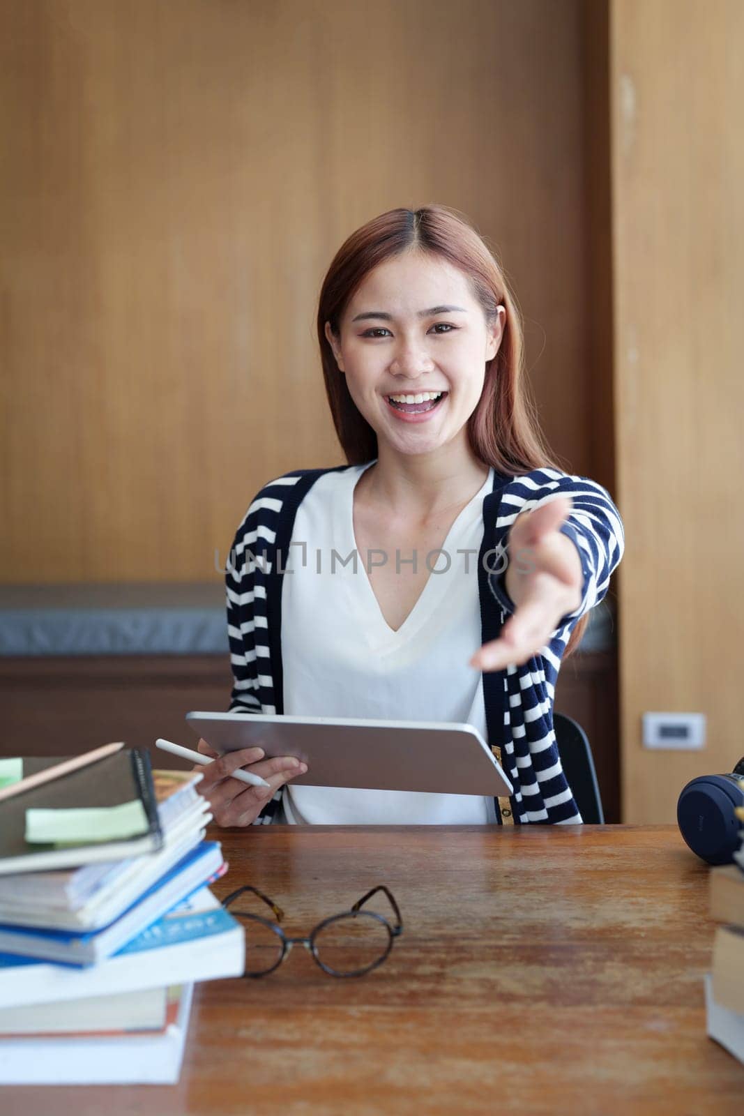A portrait of a young Asian woman showing a smiling face and gesturing out ideas while using a tablet computer studying online at a library by Manastrong