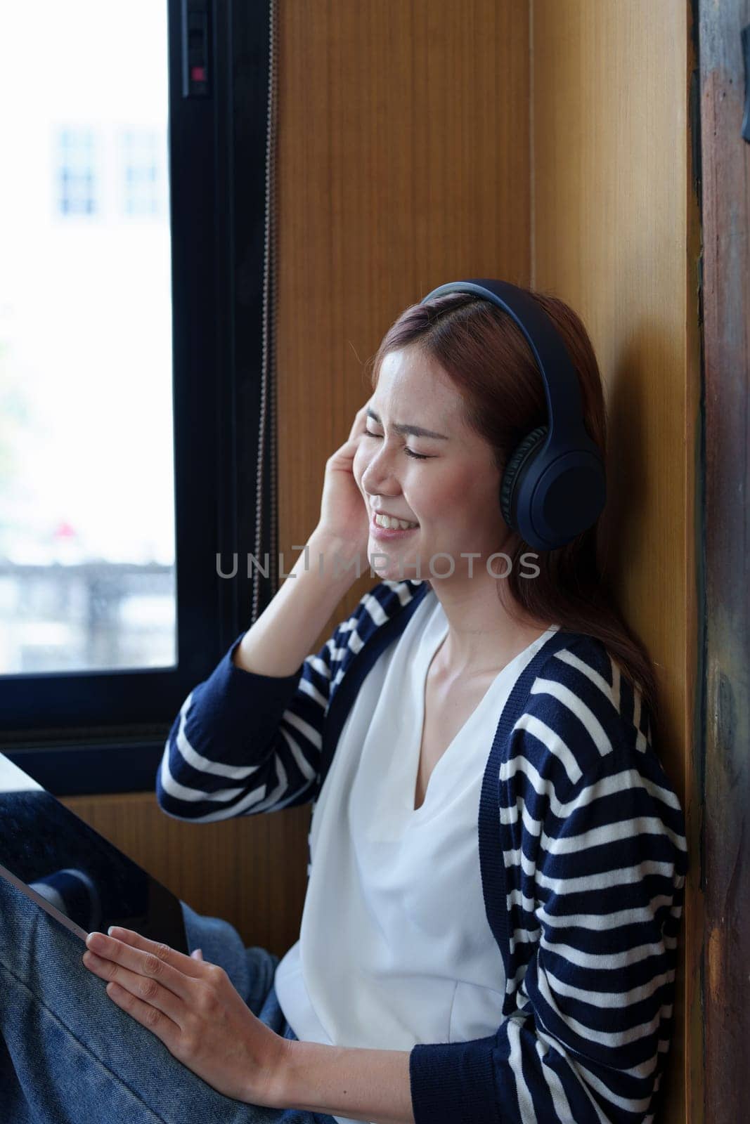 A portrait of a young Asian woman with a smiling face using a tablet computer and wearing headphones over her ears is sitting happily relaxing.
