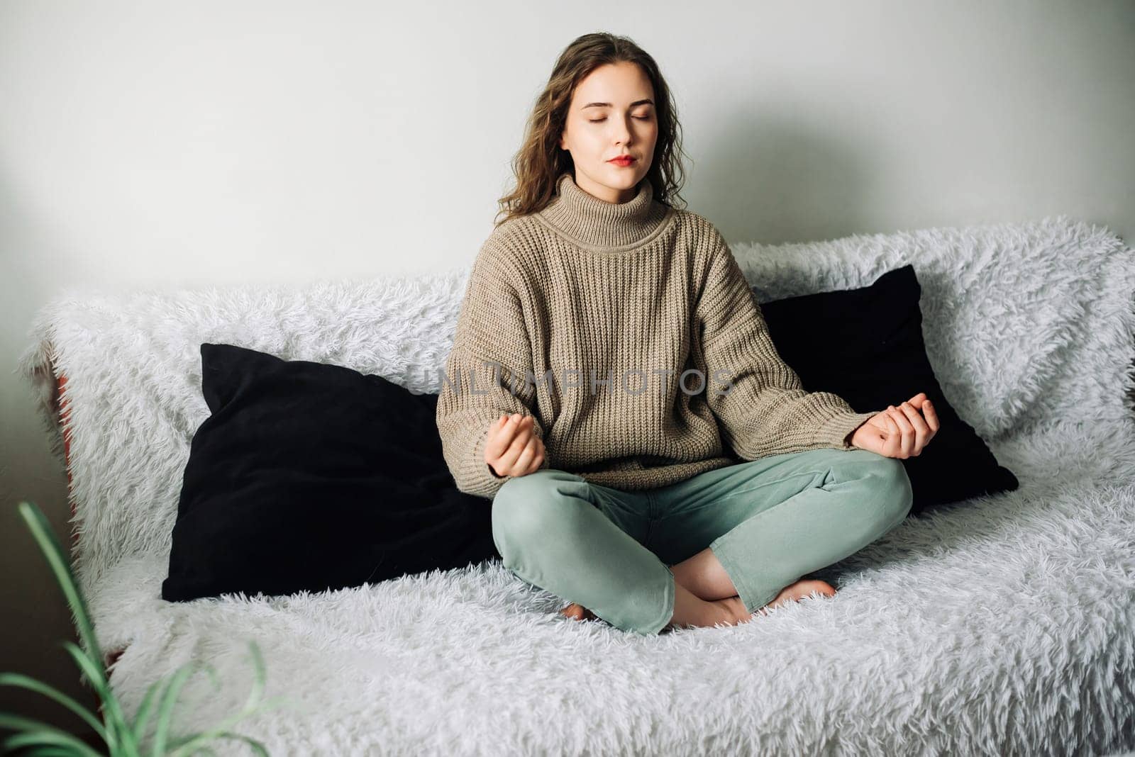 Young woman sitting in lotus position on the sofa with her hands on her knees, meditating, trying to relax her mind and relieve stress.