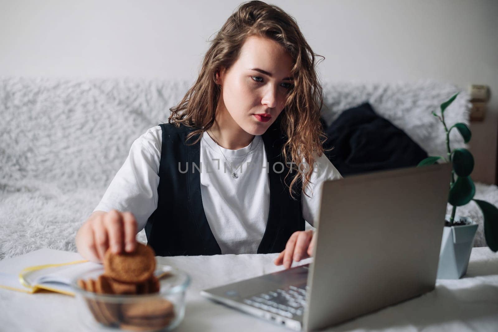 Young woman watching series or podcasts, looking at the screen with interest, holding cookies in hand, sitting at the table with a glass jar and laptop, smiling happily. Unconscious Eating Disorder.