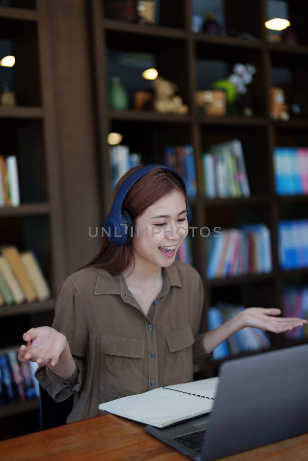Portrait of a smiling Asian teenage girl wearing headphones and using a computer for online video conferencing in a library by Manastrong