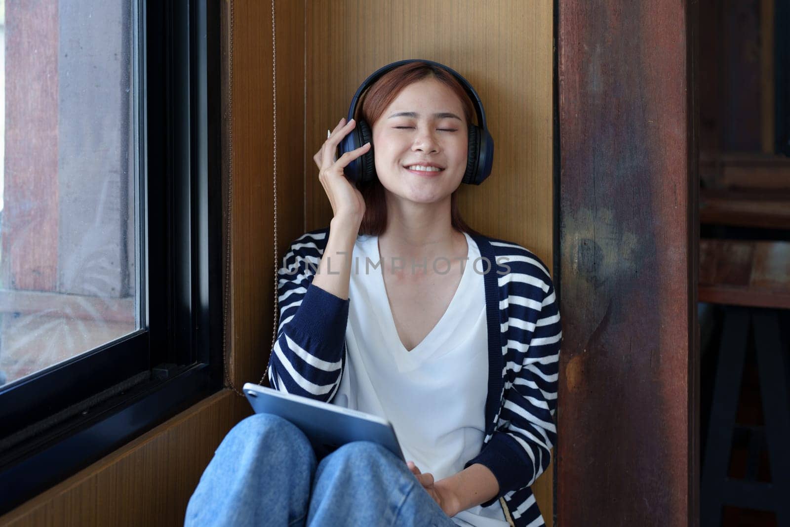 A portrait of a young Asian woman with a smiling face using a tablet computer and wearing headphones over her ears is sitting happily relaxing.