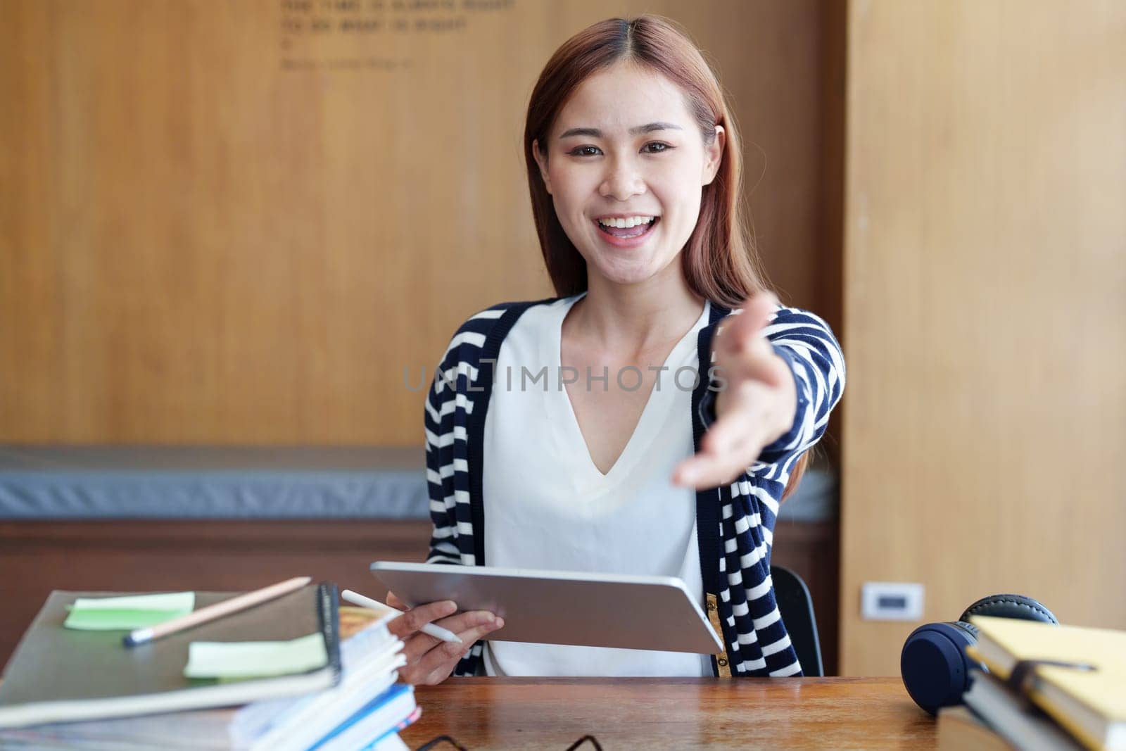A portrait of a young Asian woman showing a smiling face and gesturing out ideas while using a tablet computer studying online at a library.