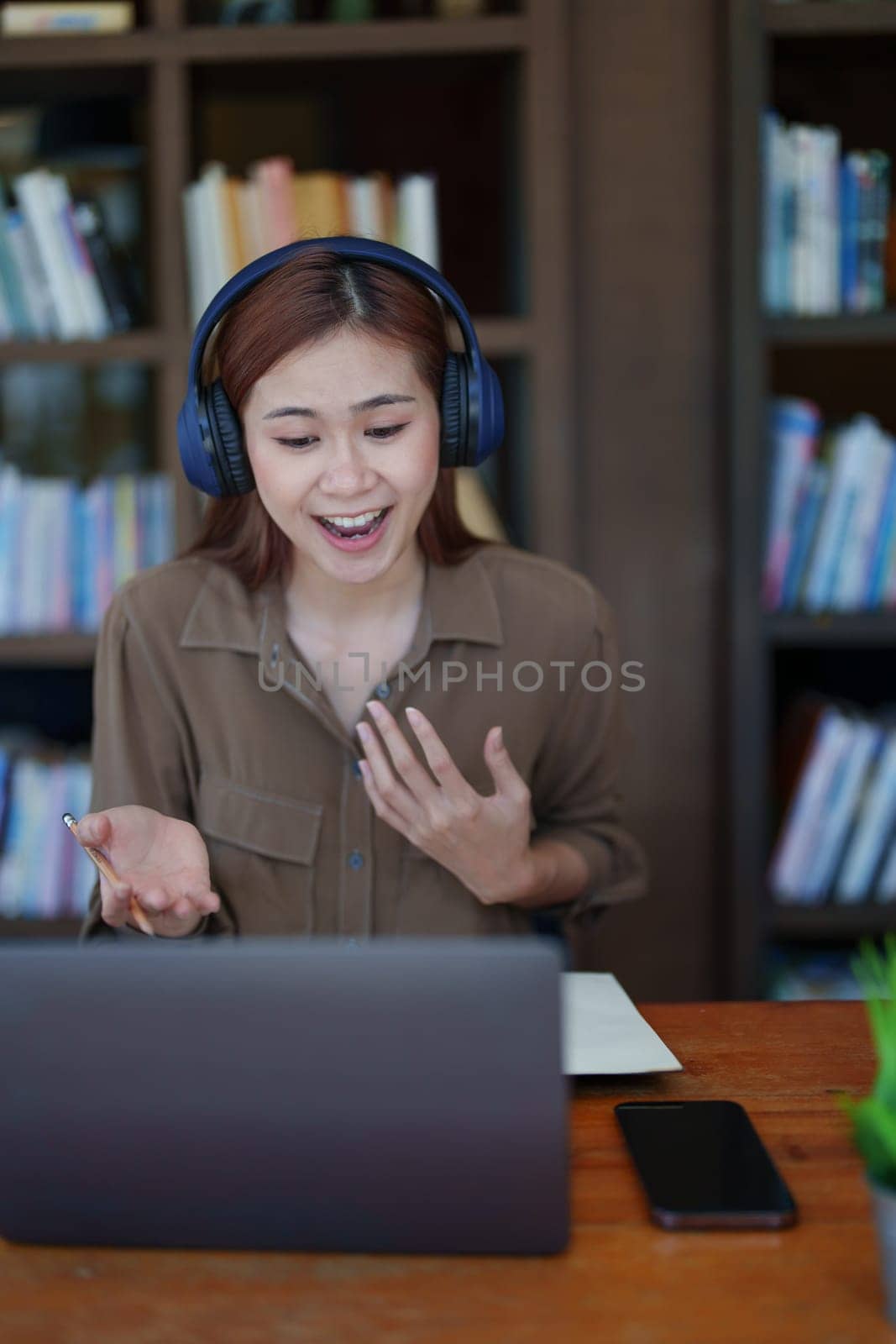 Portrait of a smiling Asian teenage girl wearing headphones and using a computer for online video conferencing in a library.