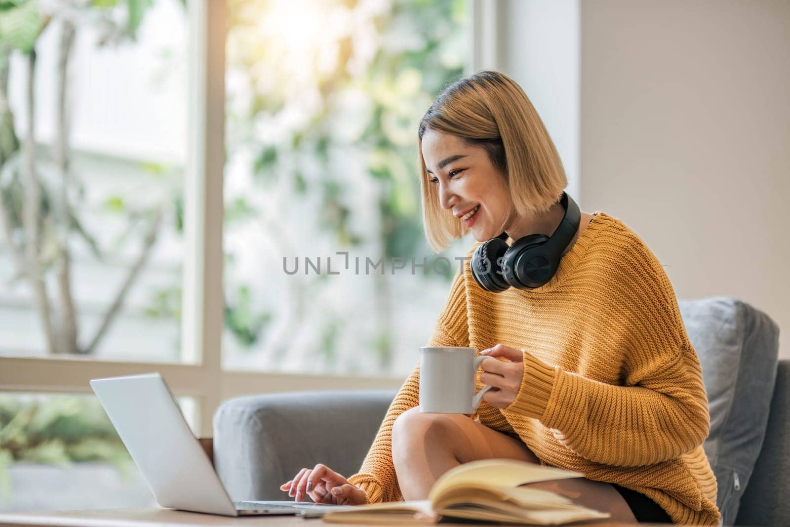 young asian woman student with headphones using laptop in a video call or online class while sitting on sofa at home.