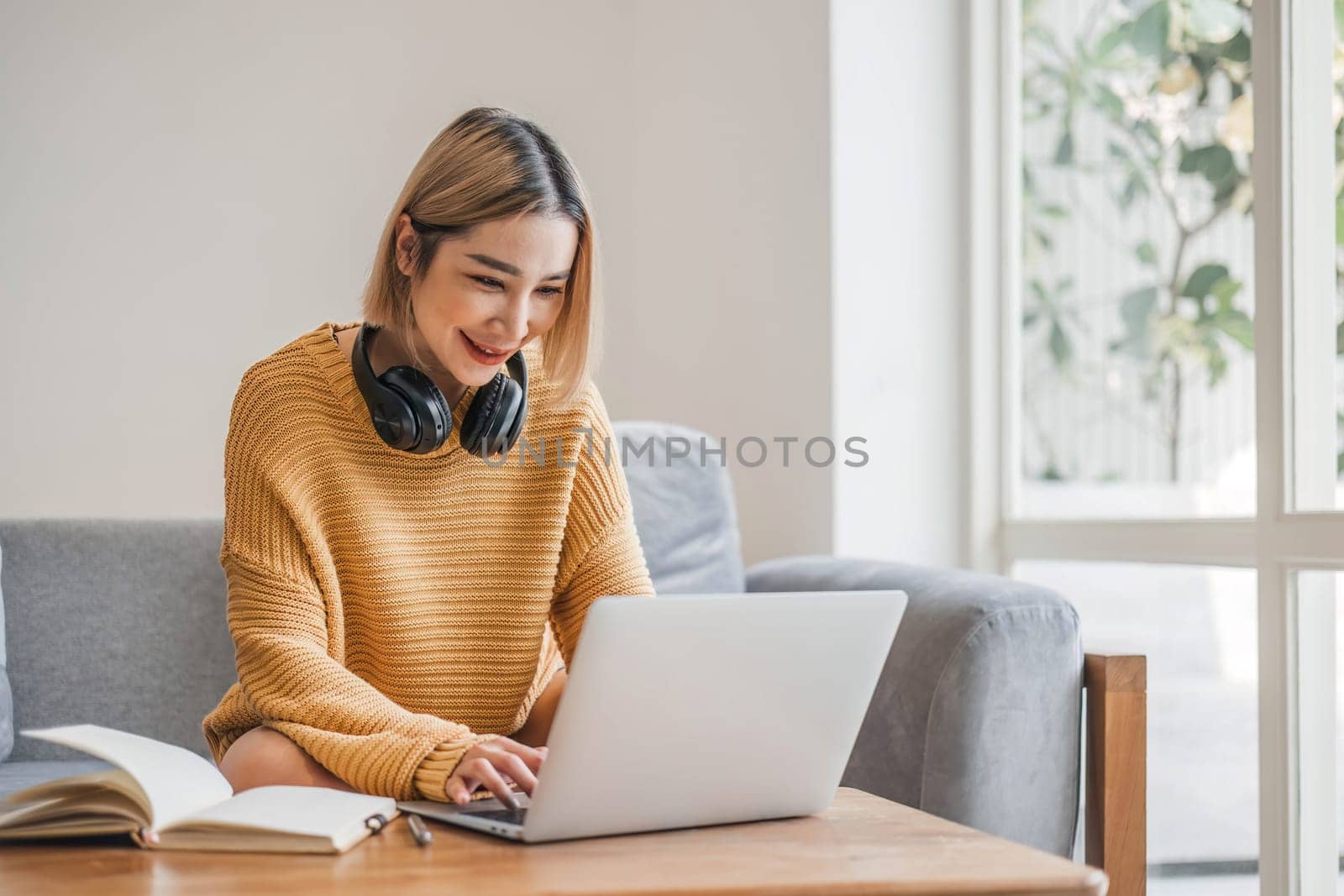 young asian woman student with headphones using laptop in a video call or online class while sitting on sofa at home.