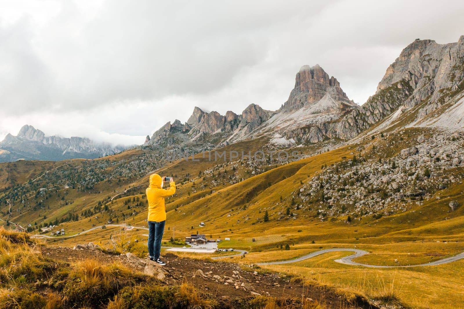 A tourist in a yellow jacket makes photos using her smartphone of meadows nestled in the picturesque Dolomites Alps.