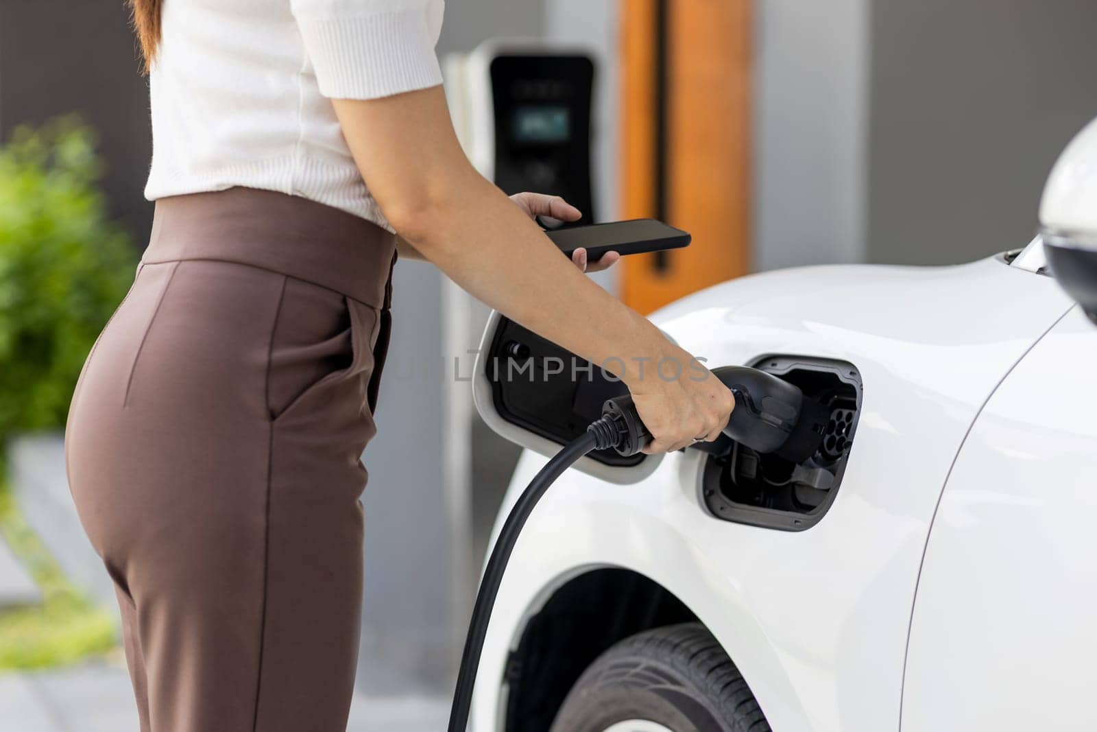 Closeup progressive woman install cable plug to her electric car with home charging station. Concept of the use of electric vehicles in a progressive lifestyle contributes to clean environment.