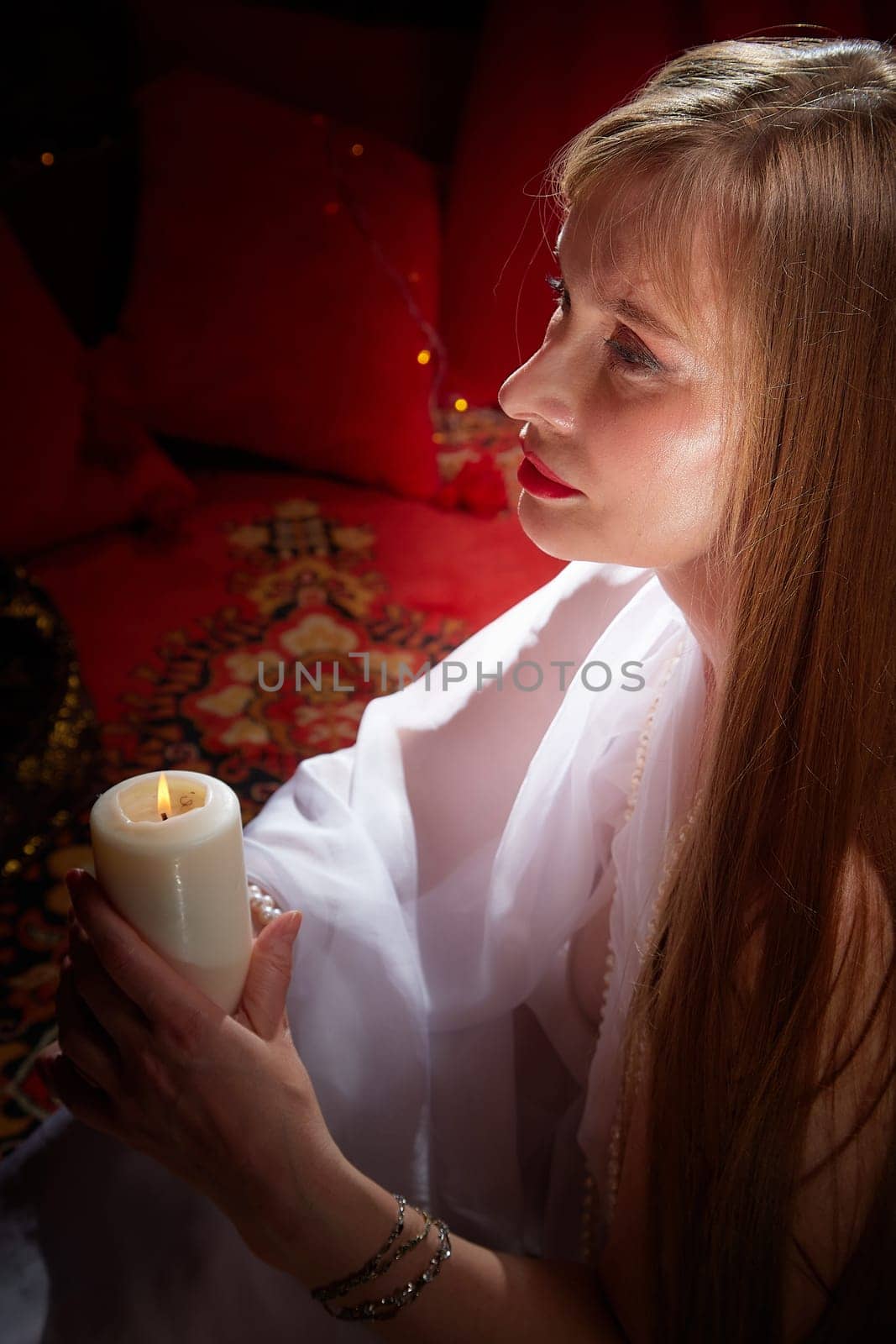 Beautiful arabian girl with candles in red room full of rich fabrics and carpets in sultan harem. Photo shoot of woman an oriental style odalisque. Model poses in sari as caring wife and hostess