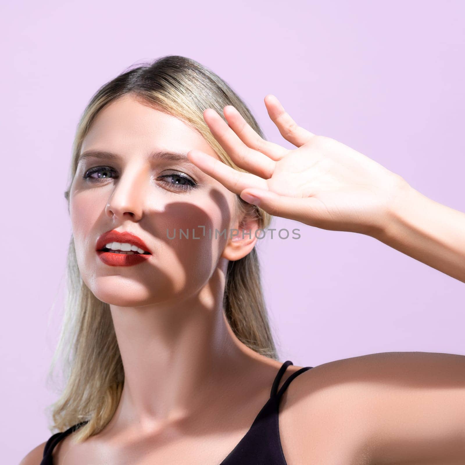 Closeup young blond hair woman with perfect skin and soft alluring facial makeup raise her hand cover her face from bright sunlight in pink isolated background for skincare sunscreen product.