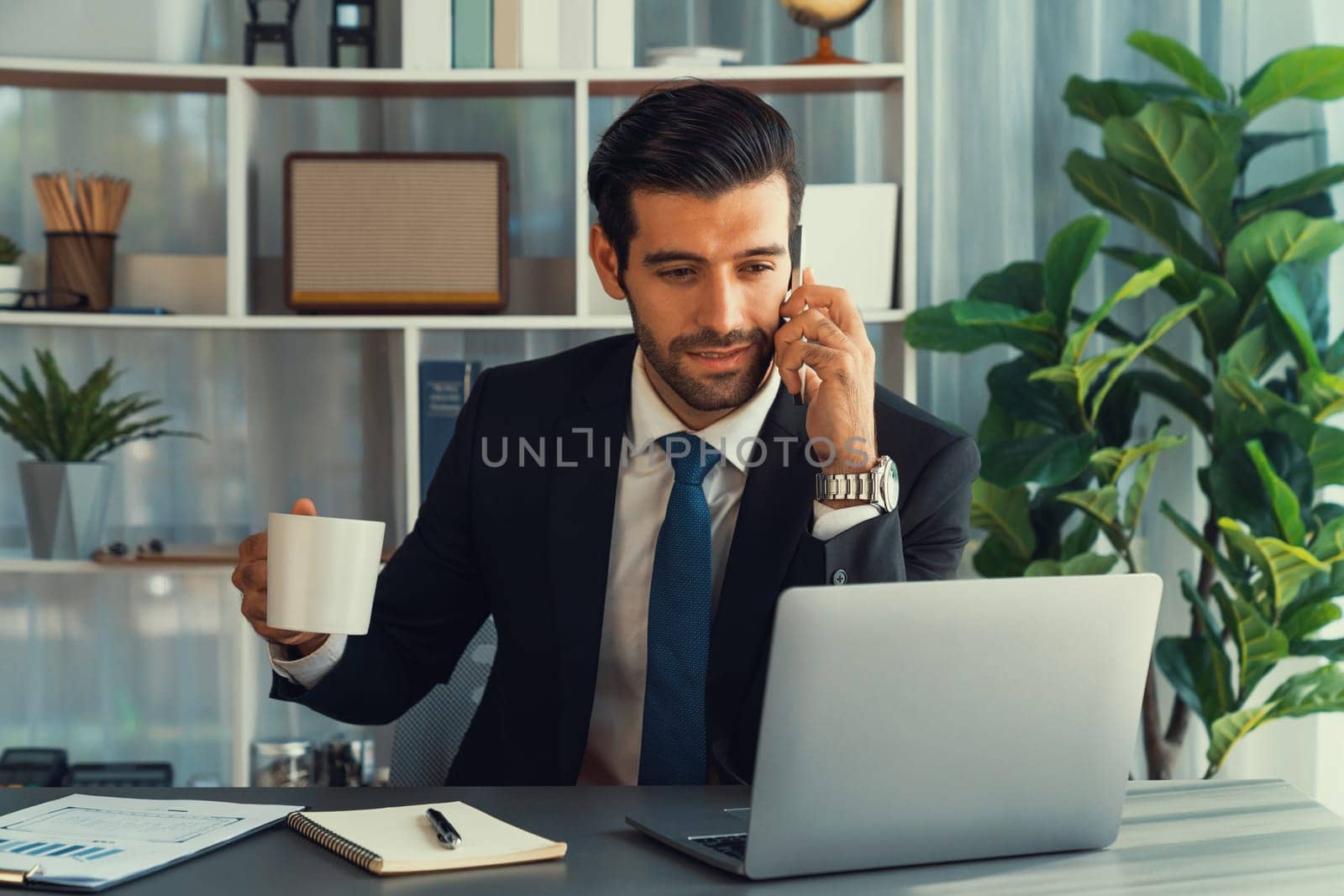 Diligent businessman busy talking on the phone call with clients while working with laptop in his office as concept of modern hardworking office worker lifestyle with mobile phone. Fervent