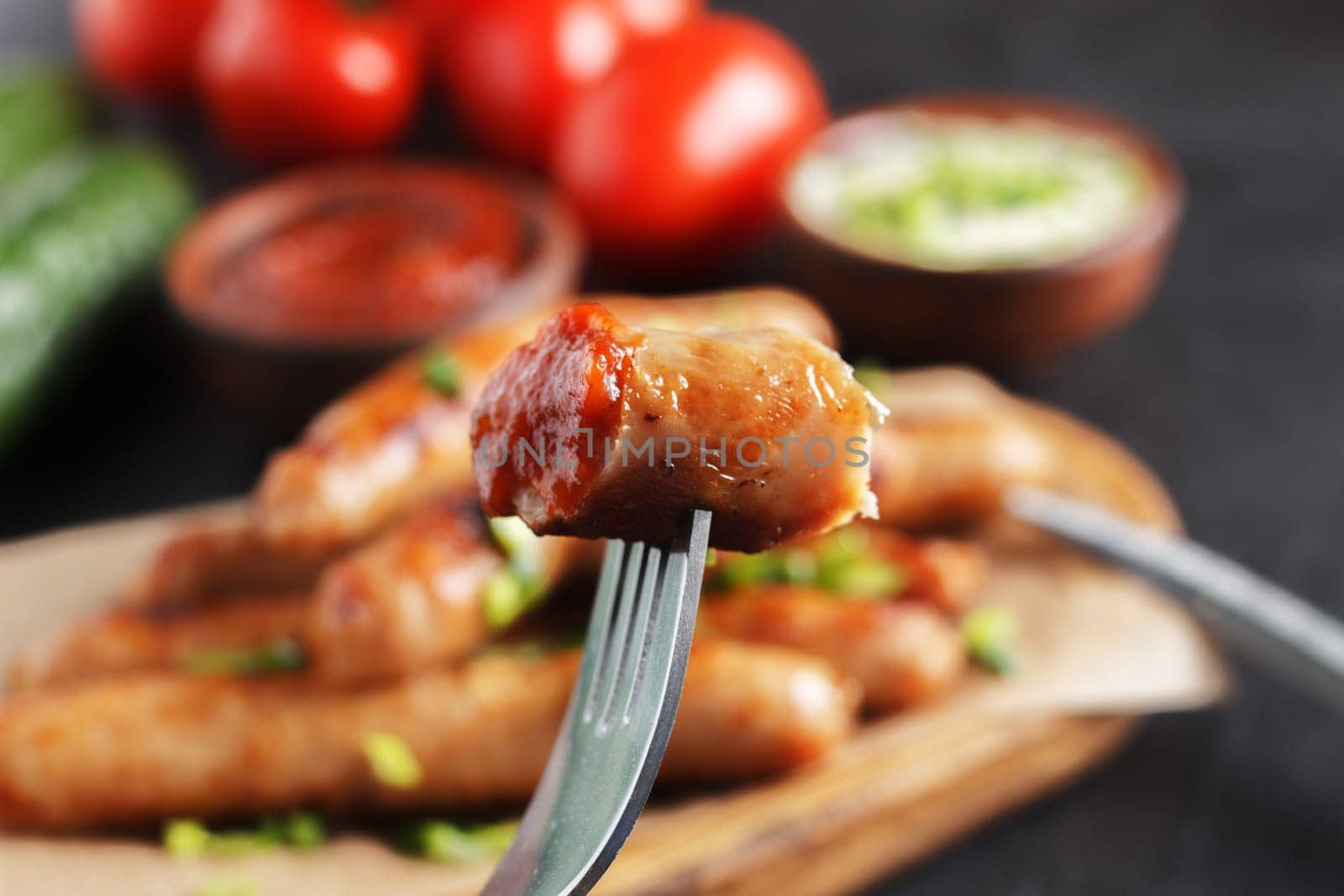 A piece of sausage on a fork against the background of sausages with vegetables and various sauces on the table.