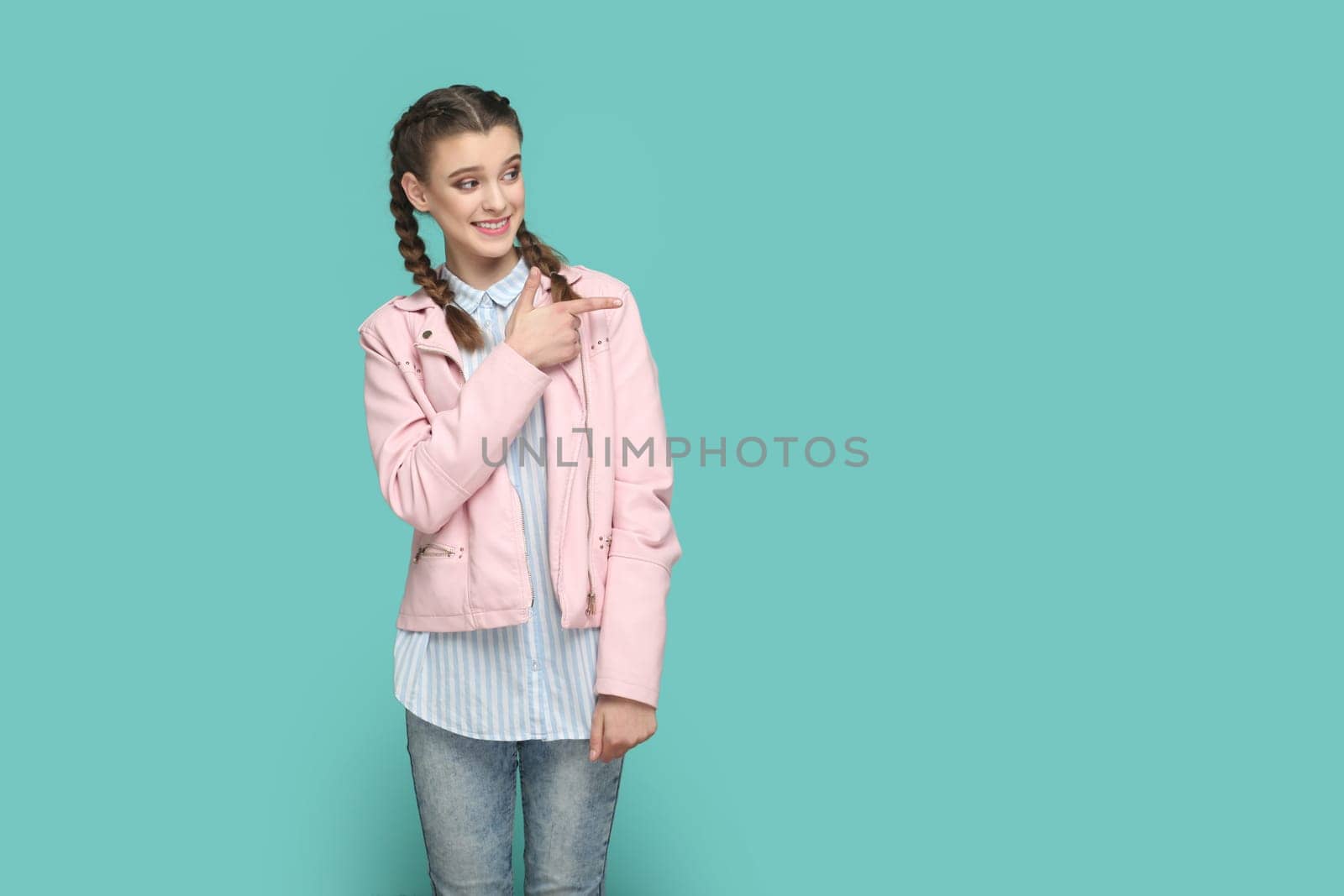 Portrait of smiling positive happy teenager girl with braids wearing pink jacket pointing aside at advertisement area, space for promotion. Indoor studio shot isolated on green background.