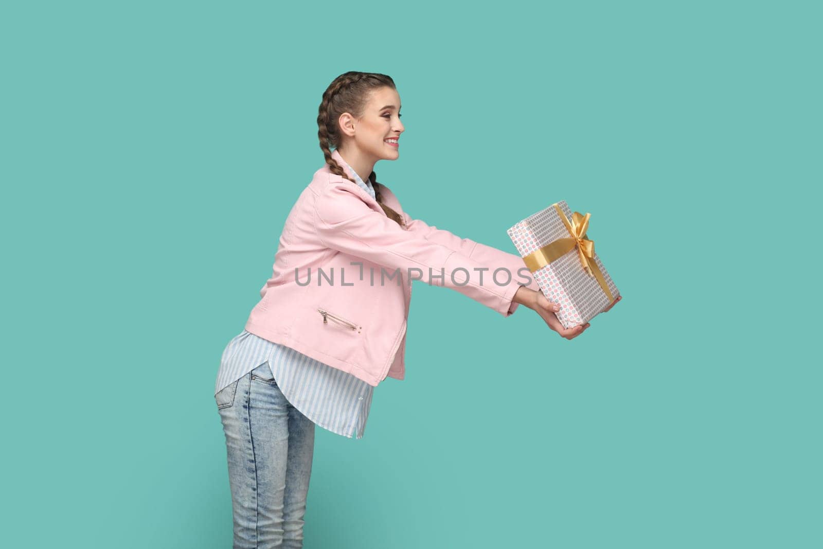 Side view portrait of excited amazed teenager girl with braids wearing pink jacket giving wrapped present box with gold ribbon, congratulating. Indoor studio shot isolated on green background.