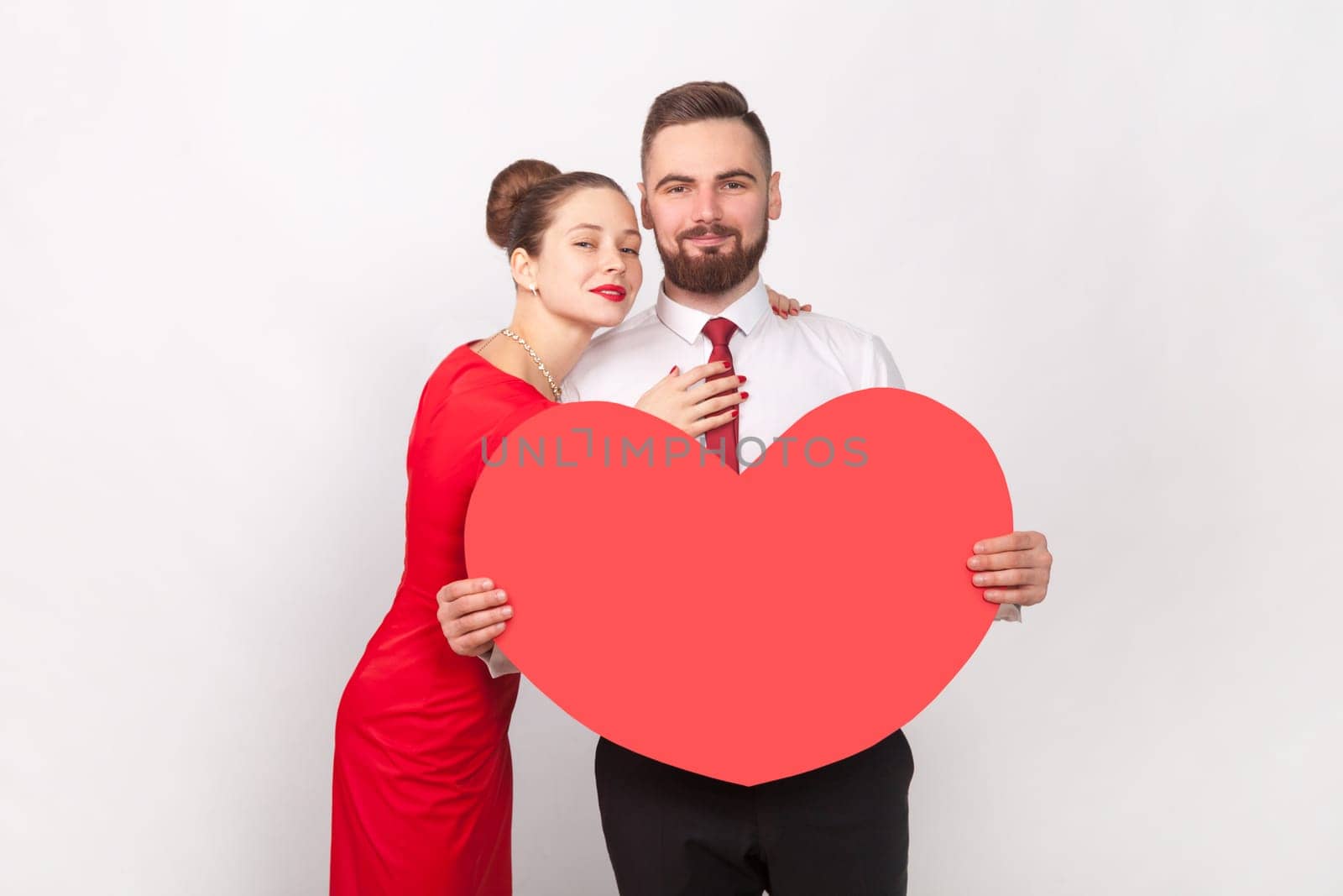 Pretty pleased couple standing together and hugging, man holding big heart, looking at camera. by Khosro1