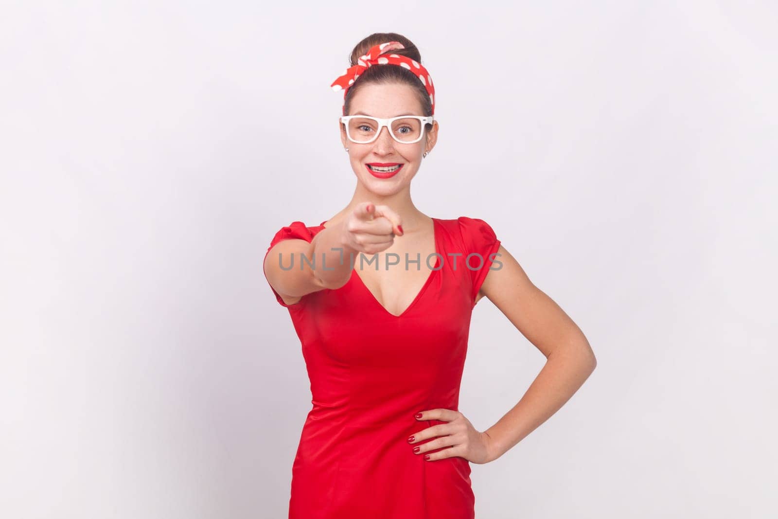 Portrait of positive woman wearing red dress and head band standing with hand on hip, pointing and choosing you, expressing optimism. Indoor studio shot isolated on gray background.