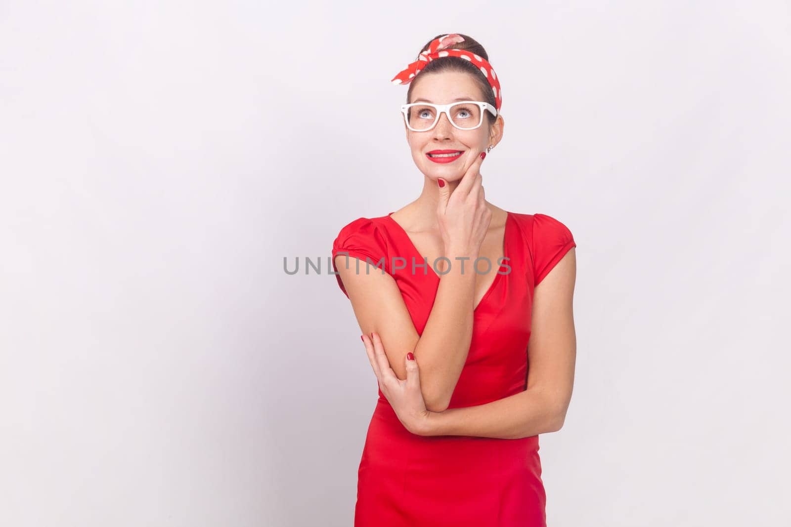 Portrait of attractive charming woman wearing red dress and head band standing looking away, dreaming about future, holding chin. Indoor studio shot isolated on gray background.