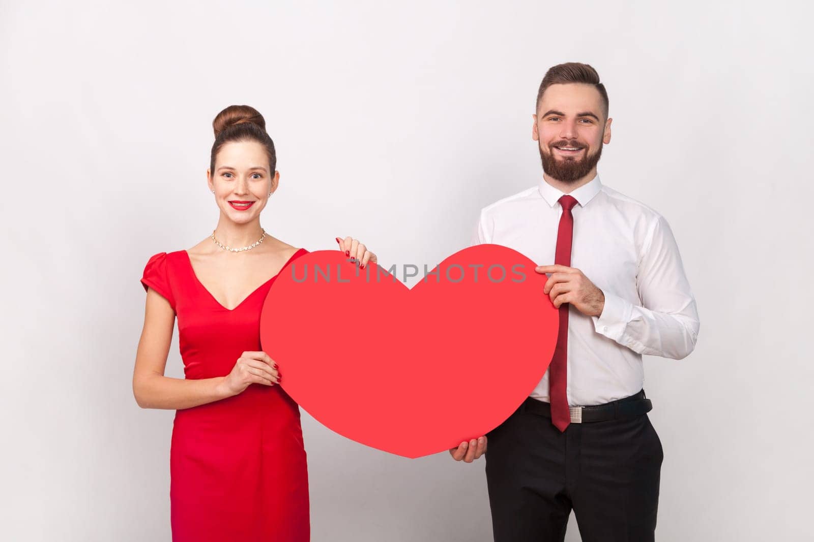 Portrait of smiling man in white shirt and woman in red dress standing together, holding big heart, celebrating Valentines Day. expressing love. Indoor studio shot isolated on gray background.