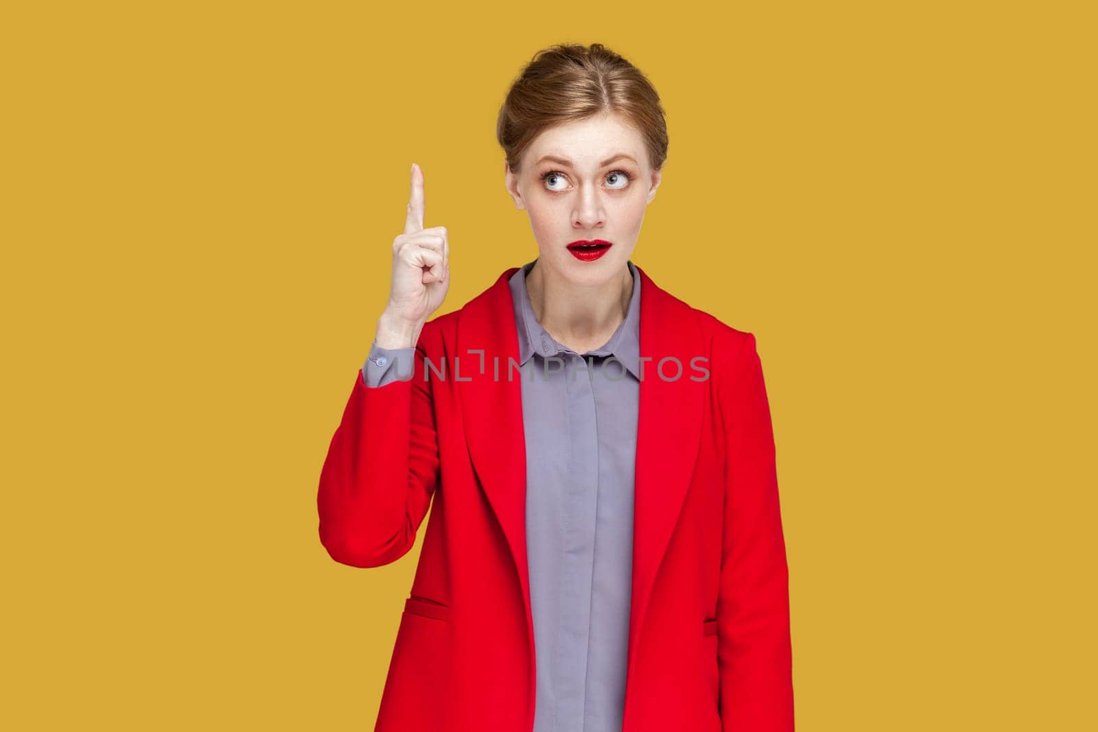 Portrait of excited amazed woman with red lips standing indicating finger upwards, having good excellent idea, wearing red jacket. Indoor studio shot isolated on yellow background.