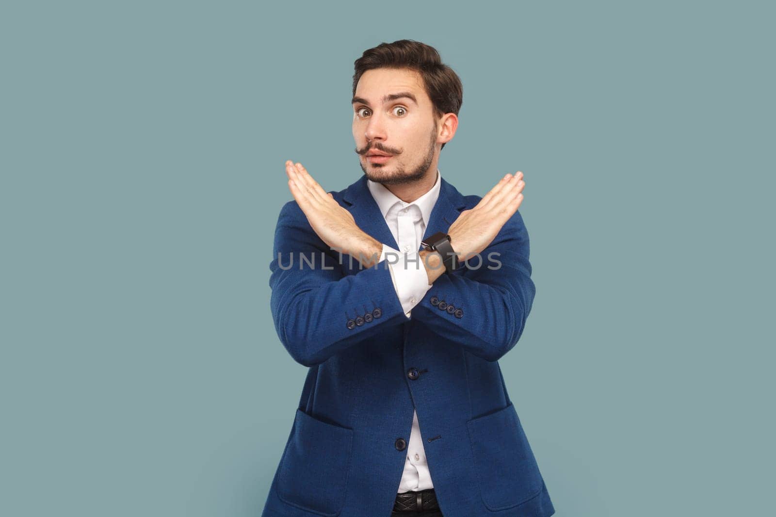 Serious strict man with mustache standing showing x sign, no way gesture, looking at camera with bossy expression, wearing official style suit. Indoor studio shot isolated on light blue background.