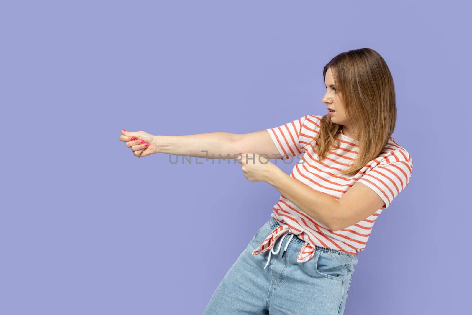 Strong ambitious woman wearing striped T-shirt pretending to pull invisible rope, concept of hard working, striving and efforts to achievements. Indoor studio shot isolated on purple background.