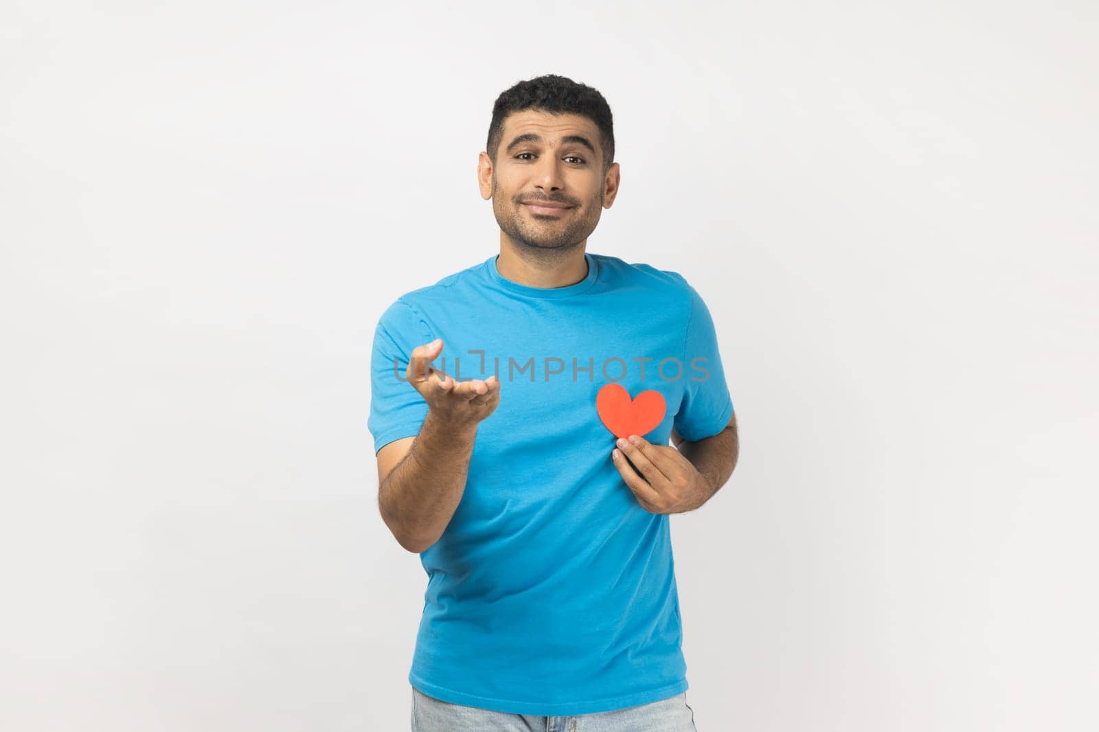 Portrait of falling in love romantic unshaven man wearing blue T- shirt standing giving his heart to his girlfriend, looking at camera, expressing gentle. Indoor studio shot isolated on gray background