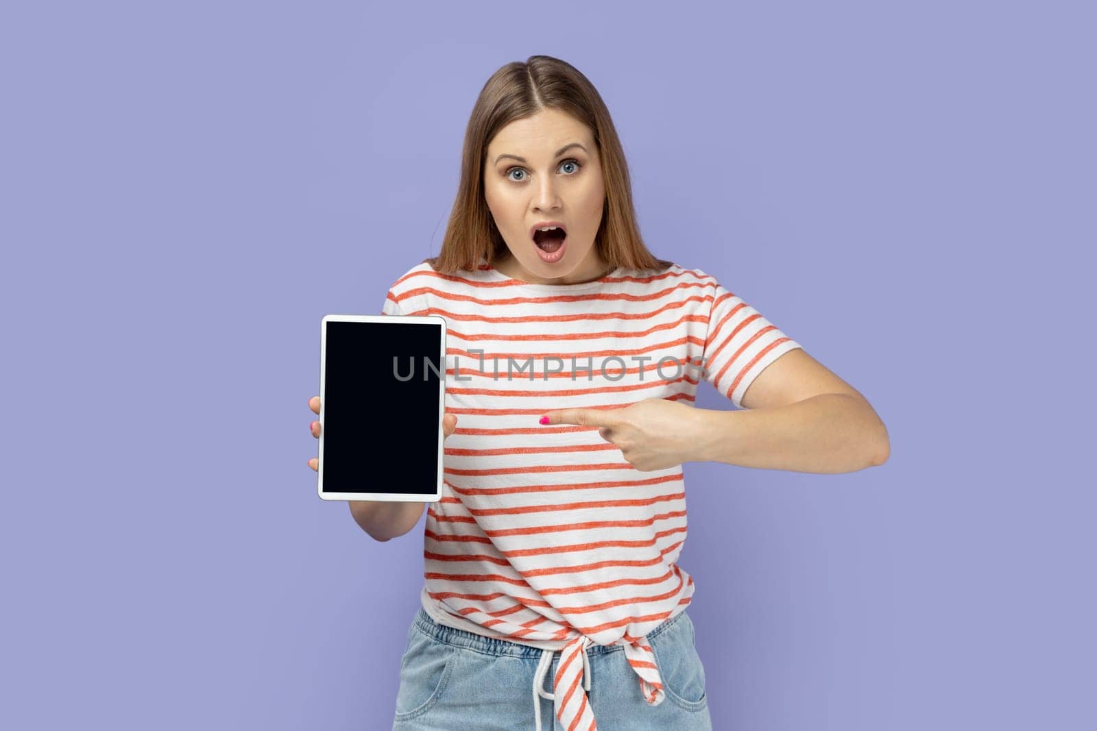 Portrait of amazed shocked surprised blond woman holding pointing at tablet with empty display, mockup screen for your advertisement. Indoor studio shot isolated on purple background.