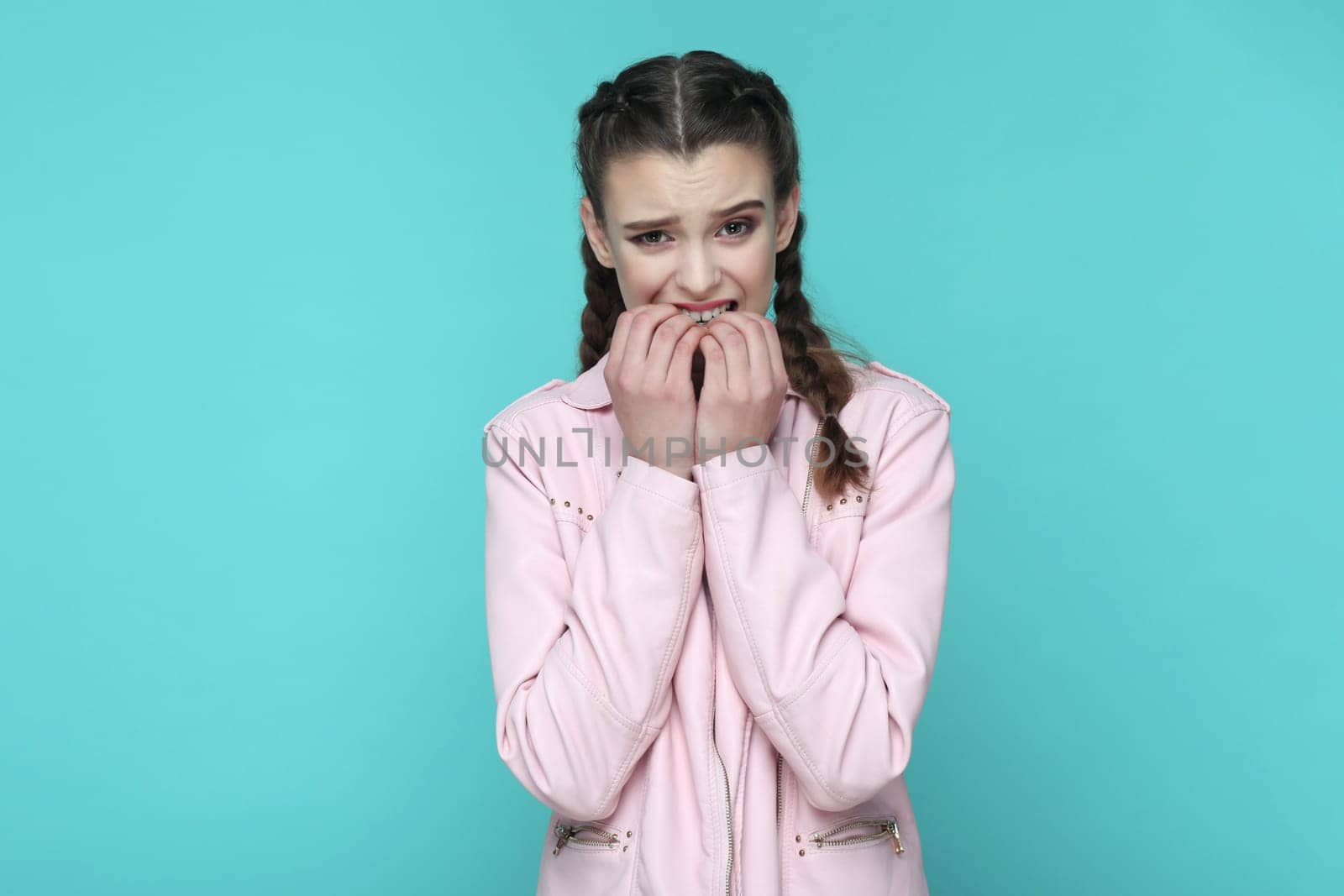 Portrait of nervous stressed teenager girl with braids wearing pink jacket being in deep depression, feels panic attack, biting her fingernails. Indoor studio shot isolated on green background.