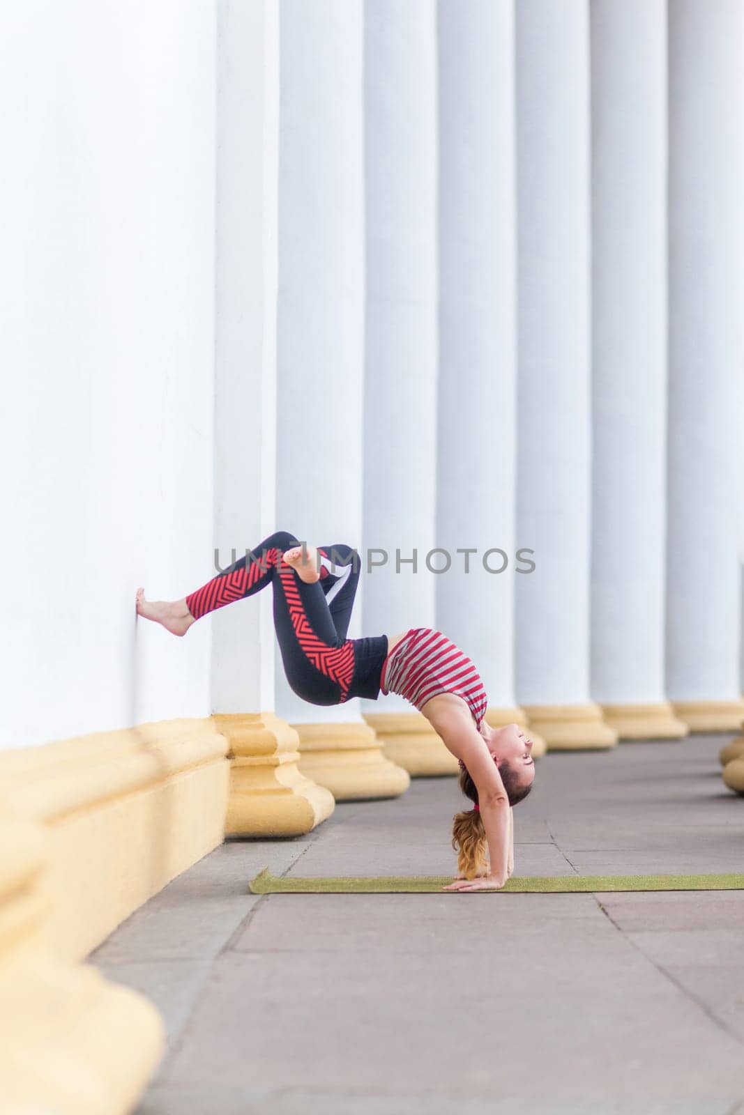 Full length side view of fit sporty strong yogi young adult woman wearing sportswear doing wall-assisted handstand pose, practicing yoga, doing pilates or fitness sport exercise outdoor.