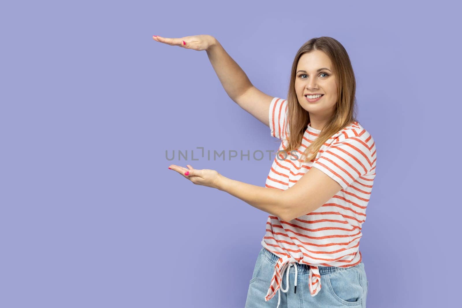 Portrait of satisfied smiling blond woman wearing striped T-shirt presenting area between hands for advertisement, showing huge size. Indoor studio shot isolated on purple background.