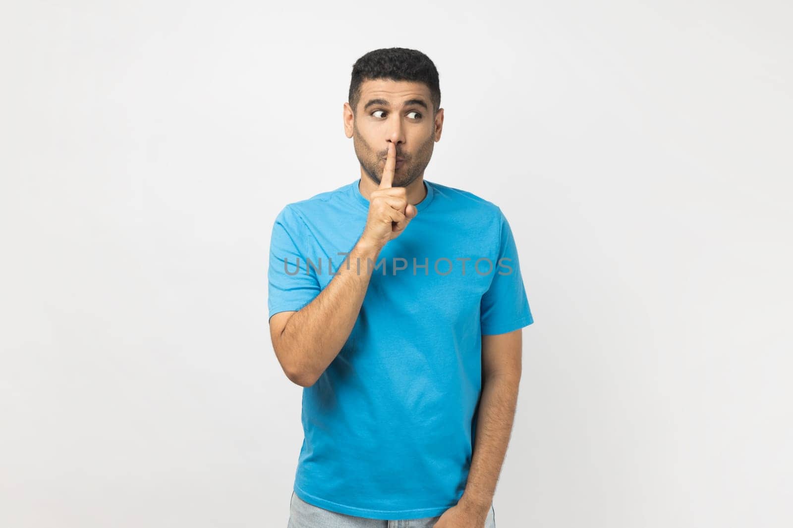 Portrait of funny positive unshaven man wearing blue T- shirt standing looking away, keeps finger near lips, promise to keep secret. Indoor studio shot isolated on gray background.
