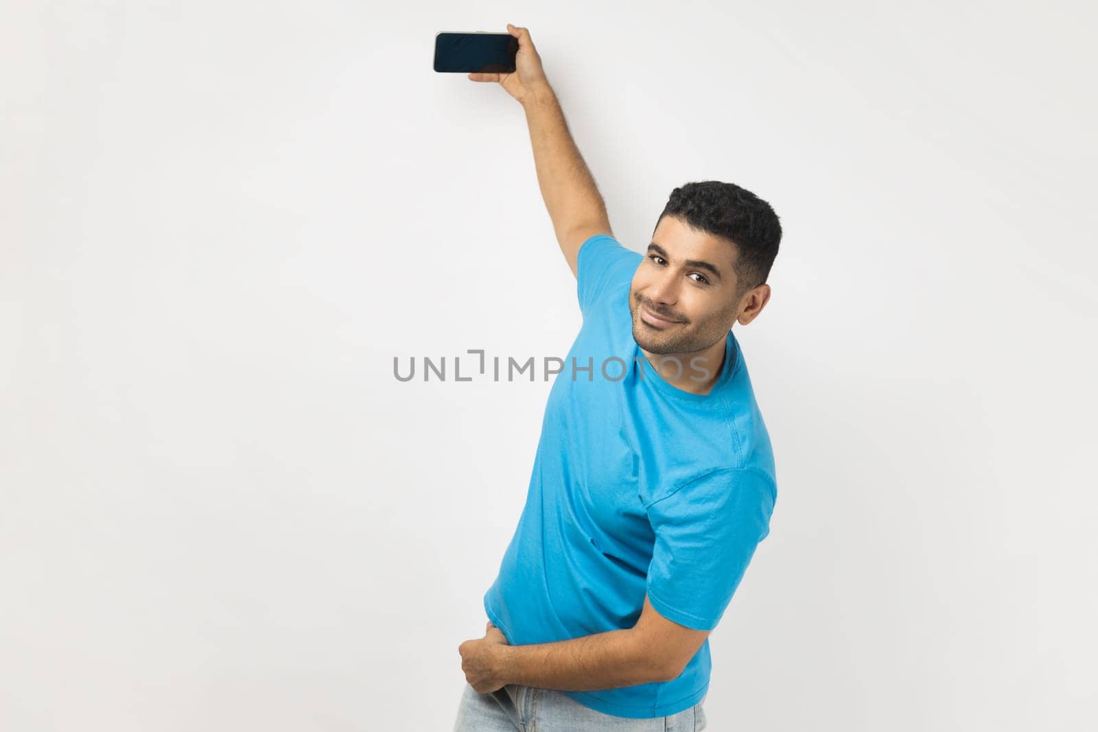 Smiling joyful man in blue T-shirt standing holding smart phone with blank screen in hands, livestreaming or having video call, looking at camera. Indoor studio shot isolated on gray background.