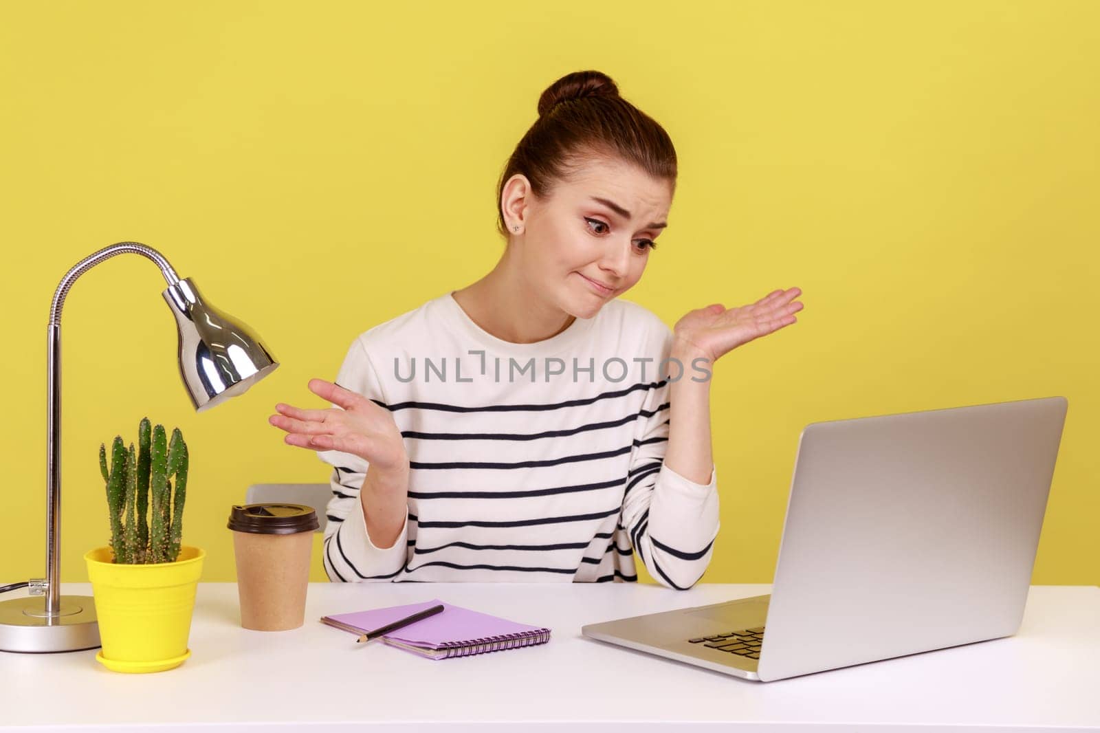 Uncertain woman office worker in striped shirt shrugging shoulders sitting on laptop at workplace, looking at monitor, having video call. Indoor studio studio shot isolated on yellow background.