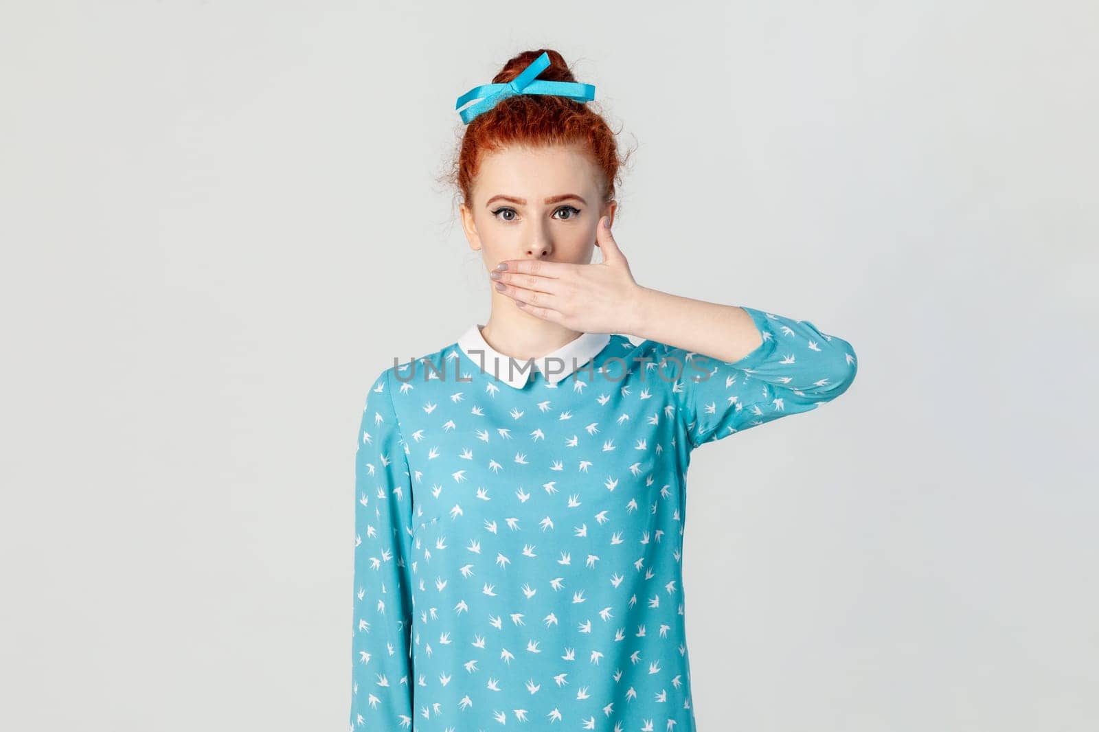 Portrait of shocked attractive ginger woman standing covering mouth with palm, promises not to say to anyone, wearing blue dress. Indoor studio shot isolated on gray background.