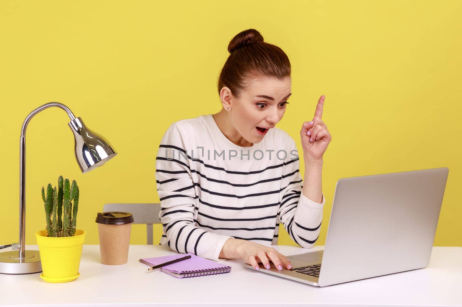 Amazed woman manager in striped shirt looking surprised by genius idea, raising finger up inspired while working on laptop. Indoor studio studio shot isolated on yellow background.