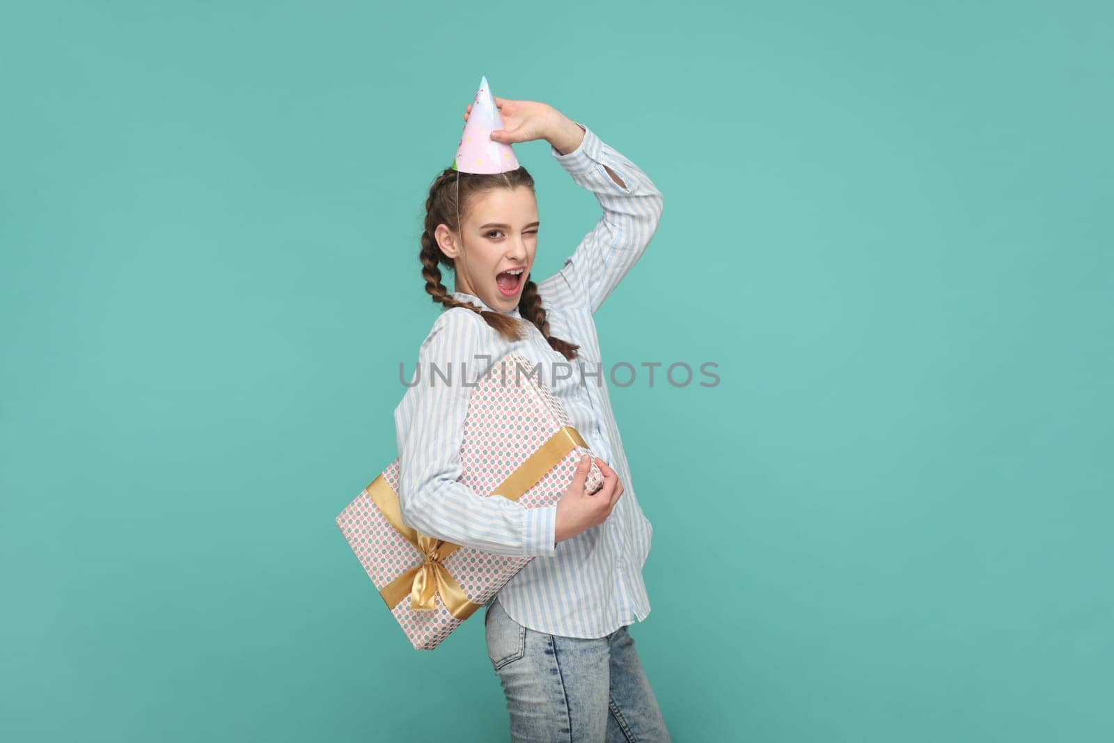 Portrait of excited cheerful teenager girl with braids wearing striped shirt and party cone, holding present box, gets gift on her birthday. Indoor studio shot isolated on green background.