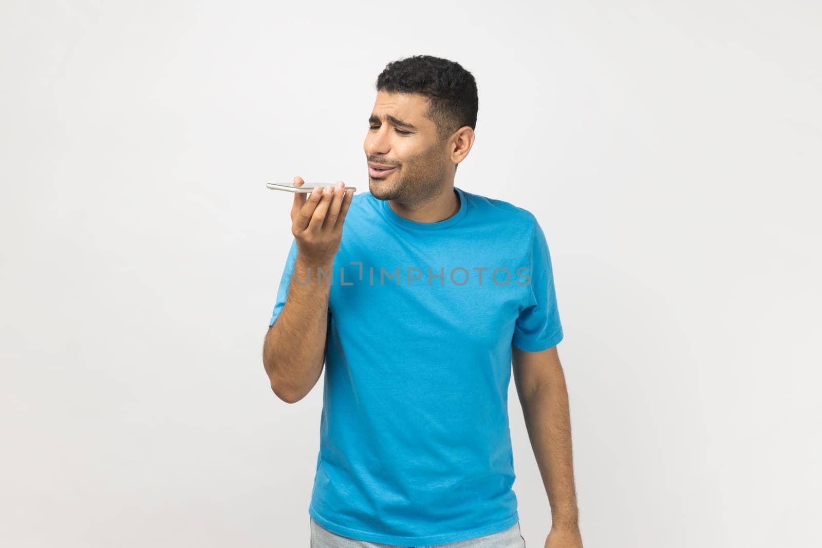 Portrait of handsome unshaven man wearing blue T- shirt standing holding smart phone, recording voice message, or using voice assistant. Indoor studio shot isolated on gray background.