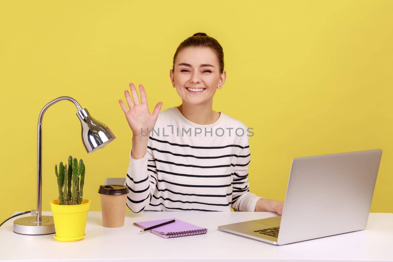 Portrait of friendly positive woman office manager waving hand to camera, showing hello gesture, looking with toothy smile. Indoor studio studio shot isolated on yellow background.
