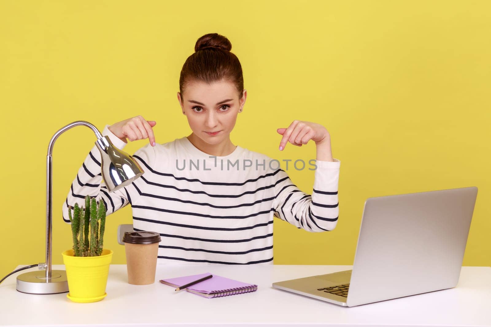 Here and right now. Young woman sitting at workplace with laptop and pointing fingers down, controlling and demanding deadline job finish. Indoor studio studio shot isolated on yellow background.