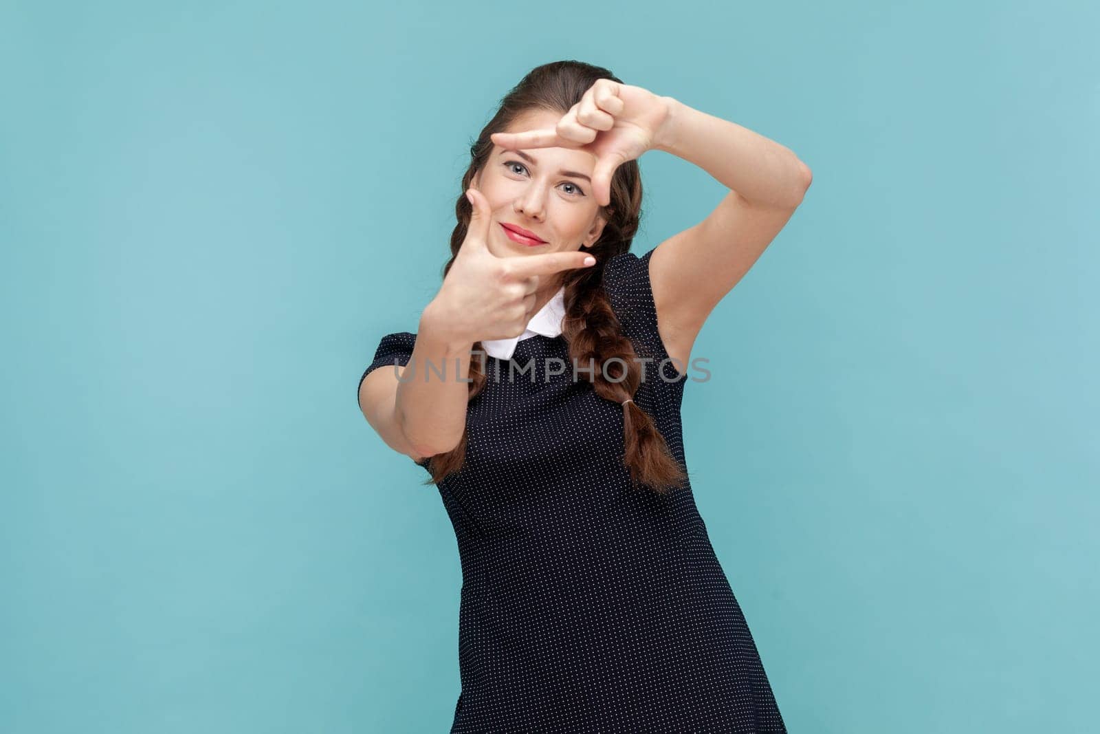 Portrait of happy ambitious woman with braids making hand frames searches perfect angle smiles broadly, wearing black dress. woman Indoor studio shot isolated on blue background.