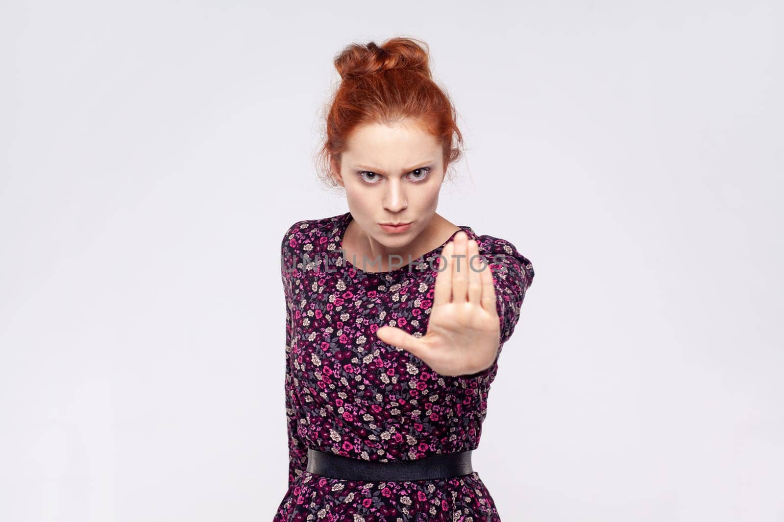 Annoyed ginger woman wearing dress making stop gesture with her palm outward, saying no, expressing denial or restriction, negative human emotions. Indoor studio shot isolated on gray background.