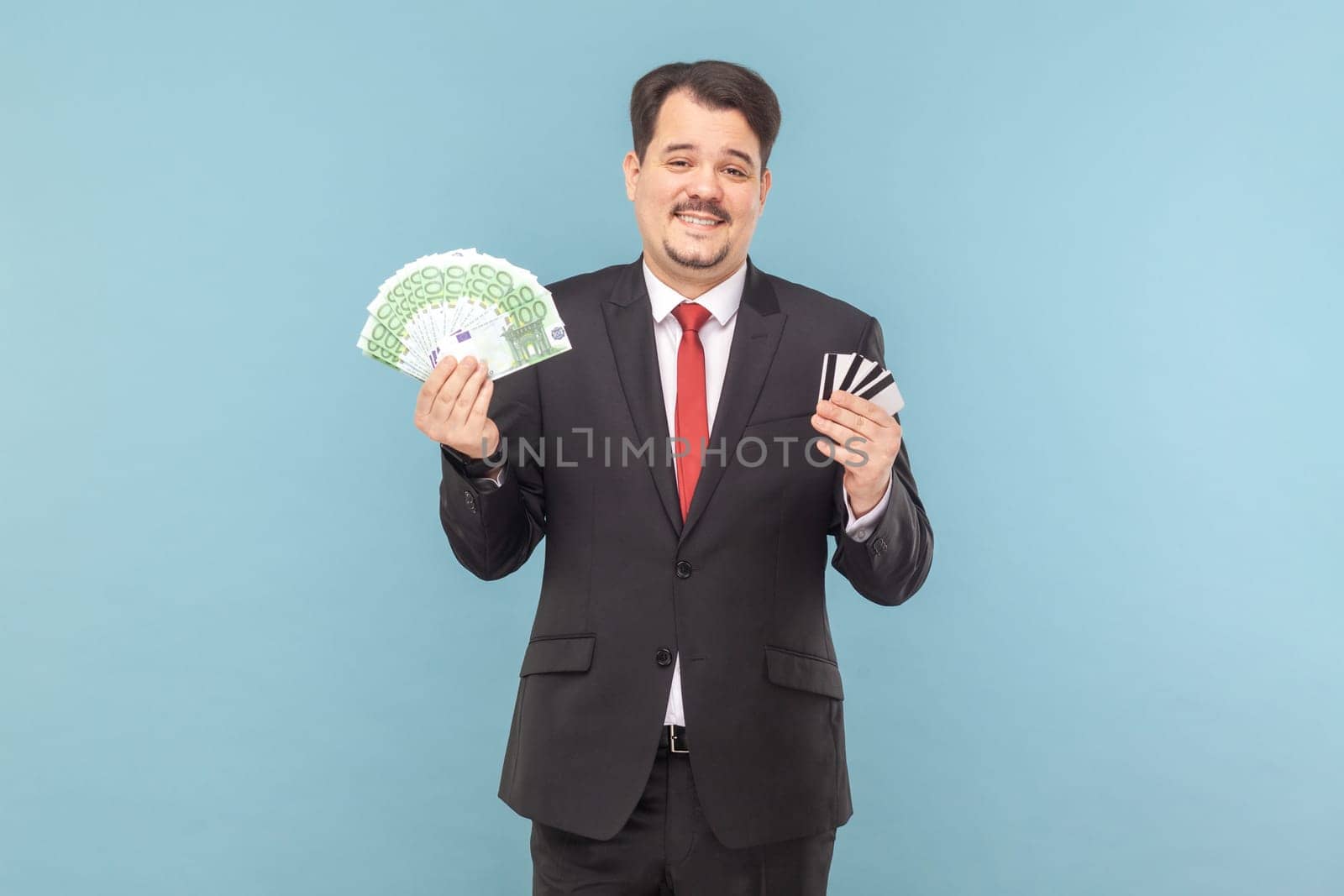 Satisfied rich man with mustache standing holding euro banknotes and credit cards in hands, being happy, wearing black suit with red tie. Indoor studio shot isolated on light blue background.