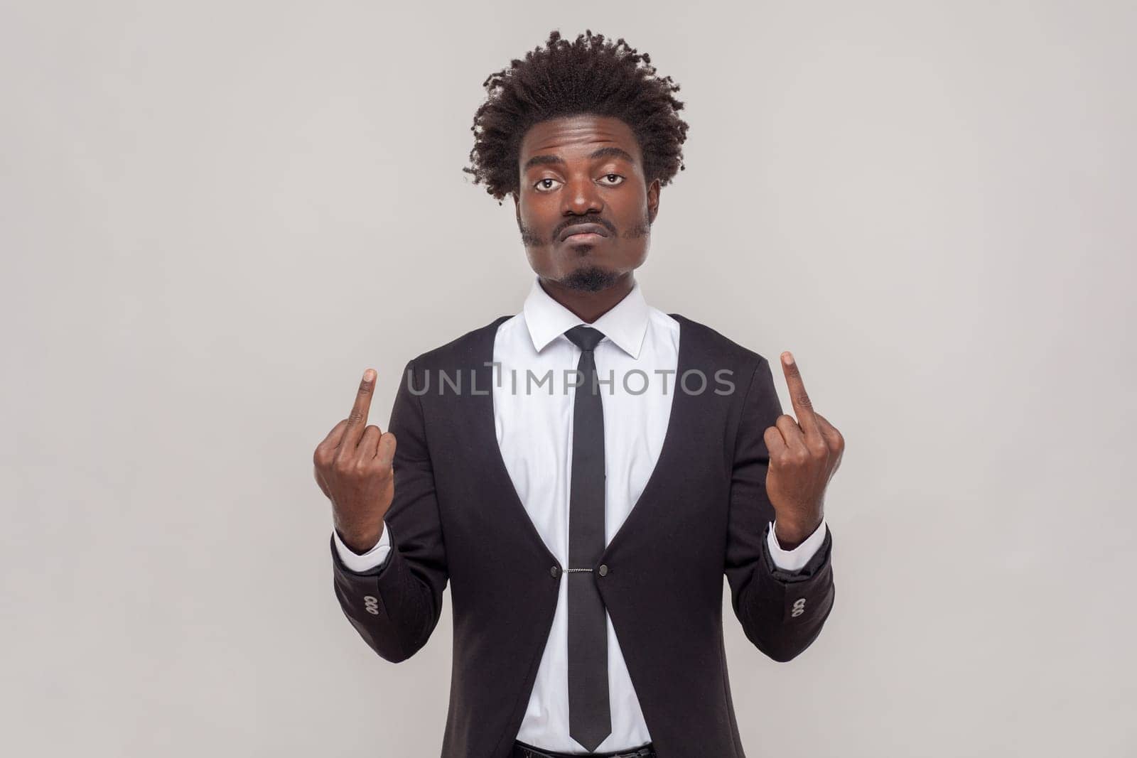 Impolite displeased man with Afro hairstyle shows middle finger, being angry with someone, purses lips frowns furious, wearing white shirt and tuxedo. Indoor studio shot isolated on gray background.