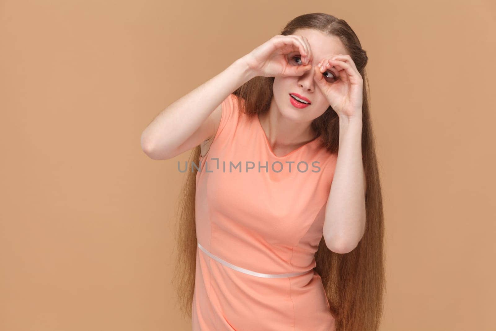 Portrait of funny surprised woman with long hair standing looking at camera through fingers, making binocular gesture, wearing elegant dress. Indoor studio shot isolated on brown background.