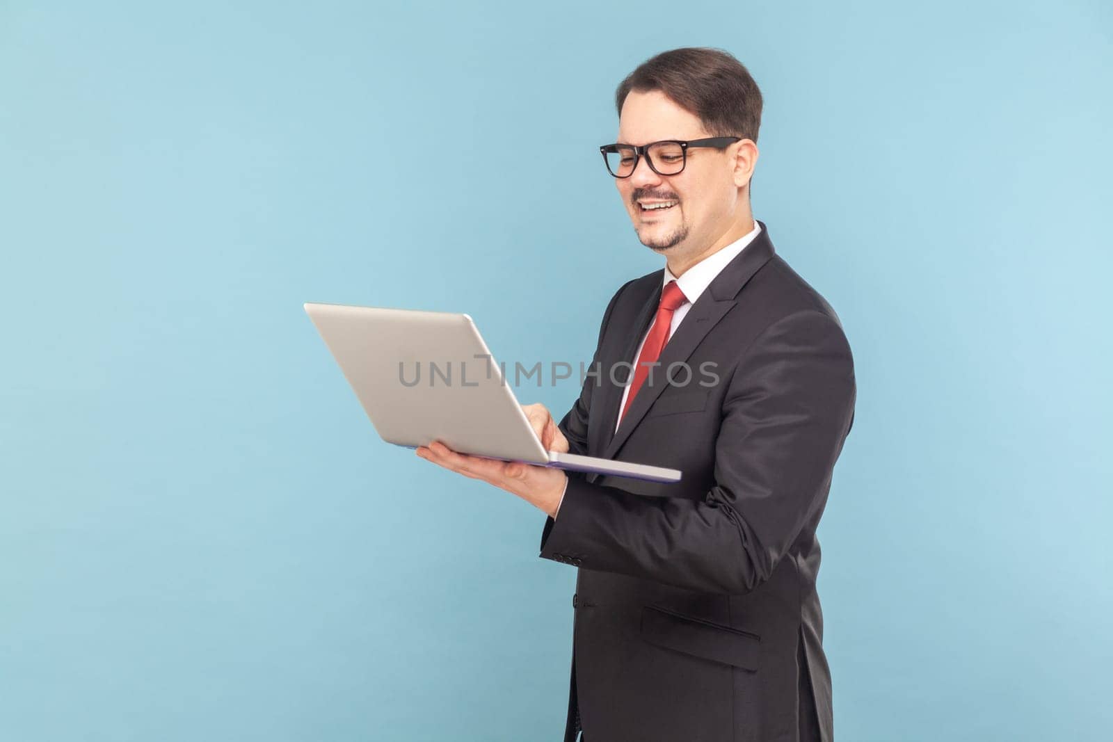 Portrait of smiling cheerful man standing and holding notebook, working on laptop online and joying his work, wearing black suit with red tie. Indoor studio shot isolated on light blue background.