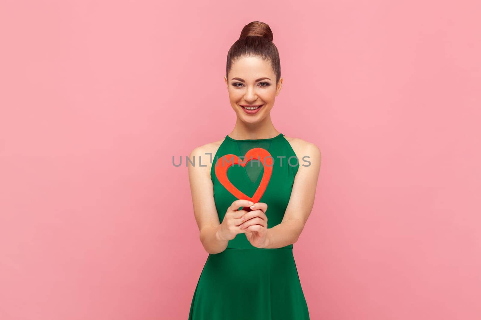 Attractive beautiful cheerful woman with bun hairstyle standing holding red heart, smiling to camera, expressing her feelings, wearing green dress. Indoor studio shot isolated on pink background.