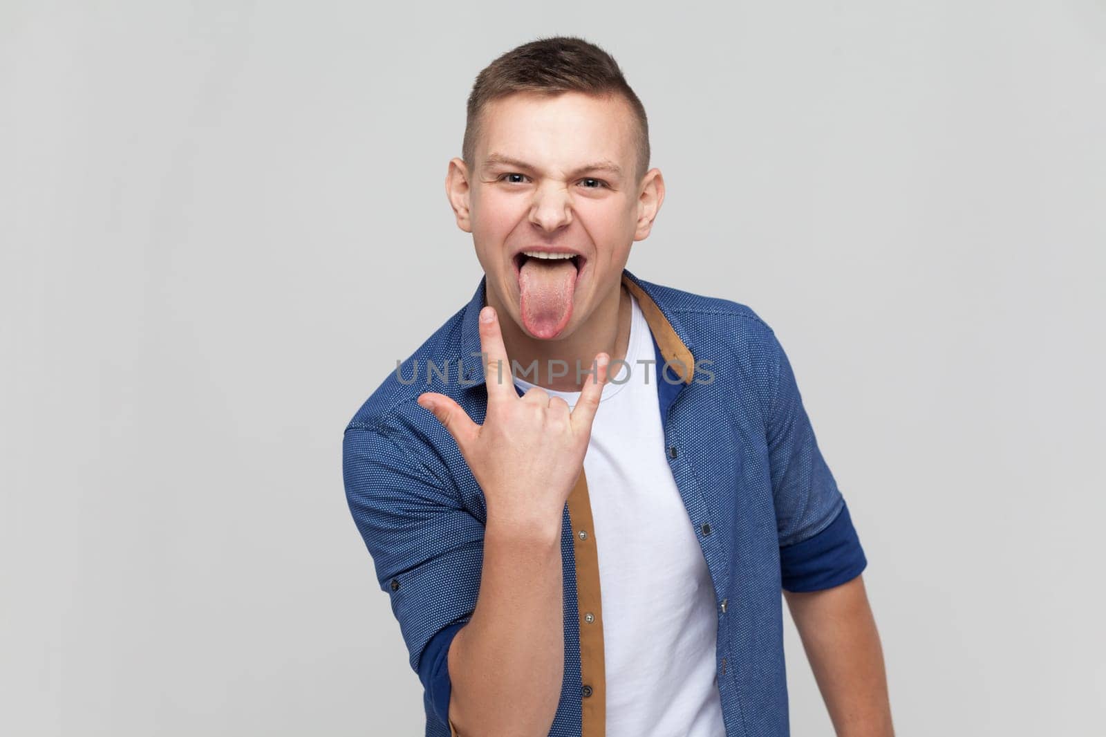 Portrait of enthusiastic crazy teenager boy in blue shirt showing rock and roll gesture and sticking out tongue, feeling to be cool rocker on festival. Indoor studio shot isolated on gray background.