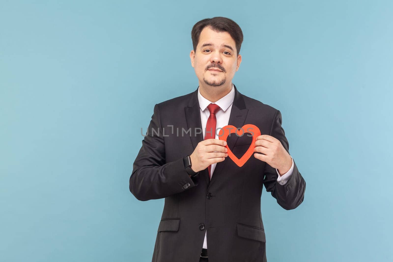 Portrait of attractive man with mustache standing holding red gesture, looking at camera with positive expression, wearing black suit with red tie. Indoor studio shot isolated on light blue background