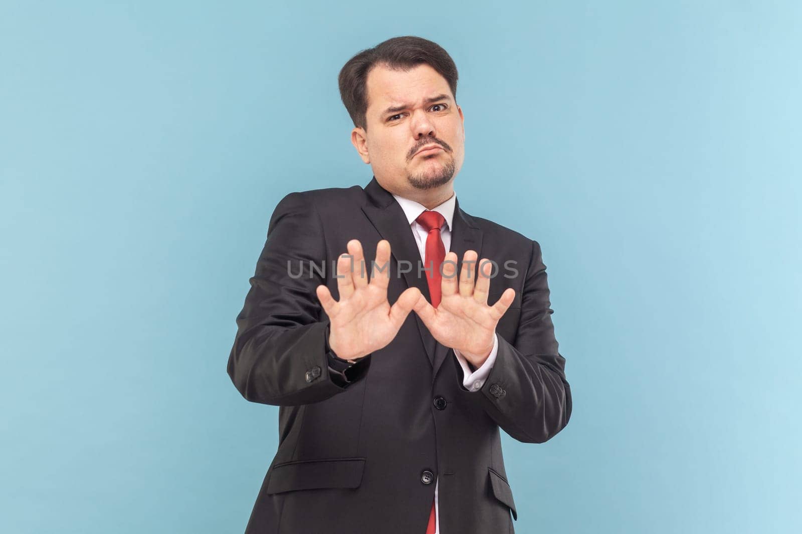 Portrait of frighten man with mustache standing raised arms, showing stop block gesture, being scared, wearing black suit with red tie. Indoor studio shot isolated on light blue background.